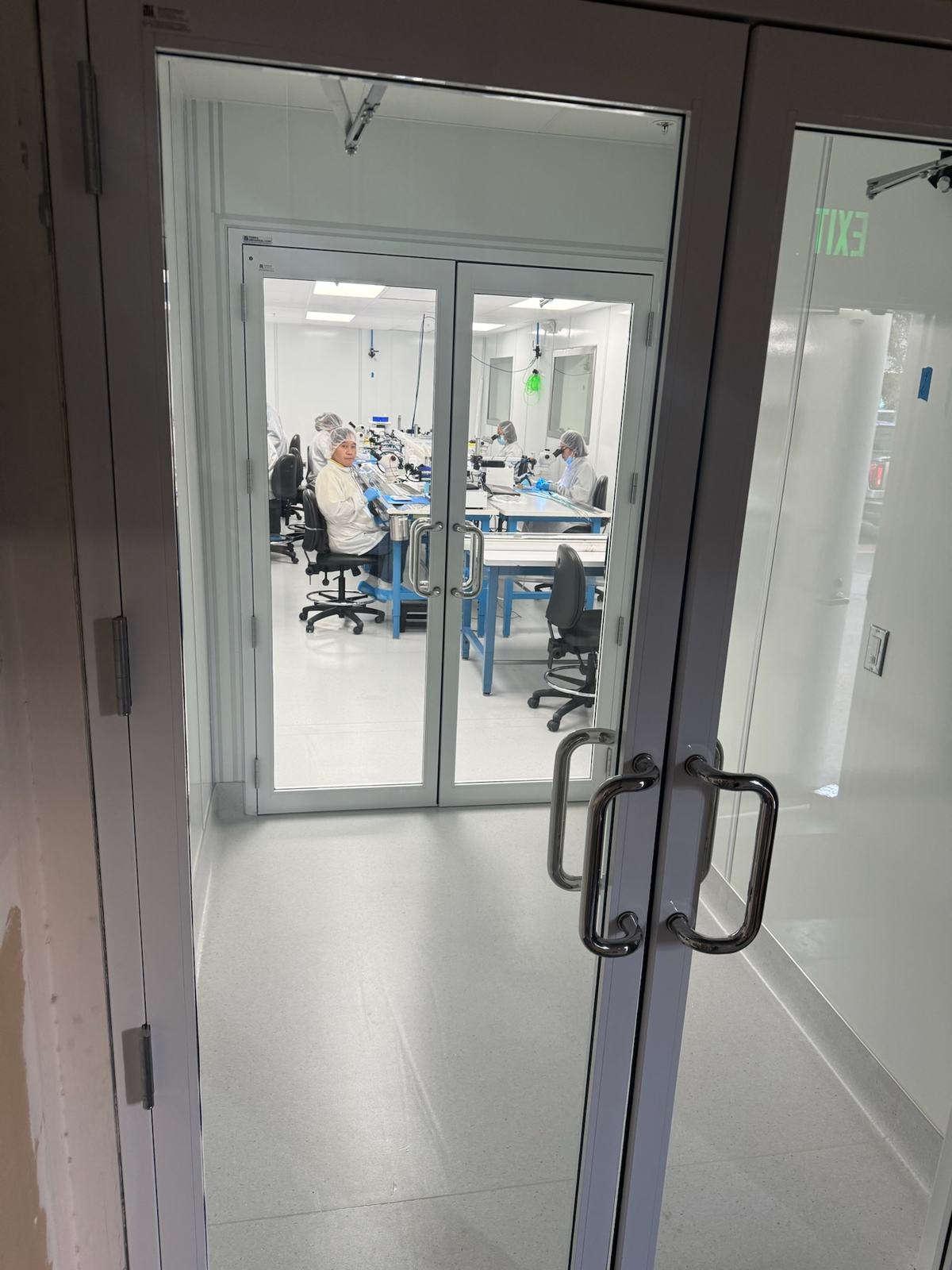 Technicians assembling catheters at microscope stations, seen through the cleanroom doors